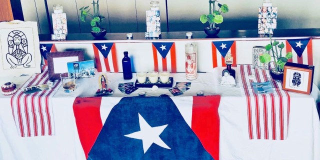 Honoring the Pulse Nightclub shooting on its anniversary: A home photograph of an altar built in a living room with a collection of white table cloth, candles, and Puerto Rican flags.