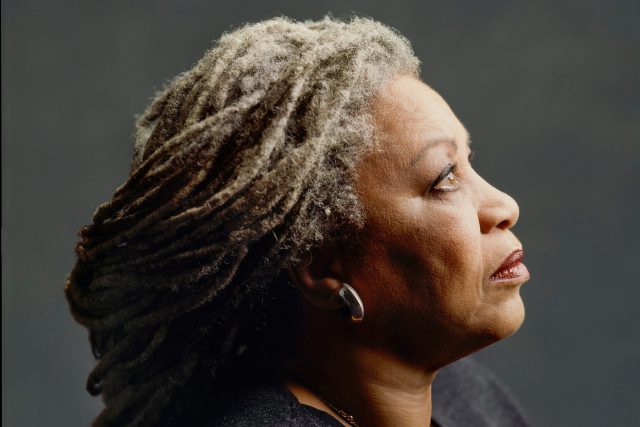 An iconic and famous portrait of Toni Morrison, looking off camera, with her grey dreadlocks pulled into a low bun just past her shoulders.