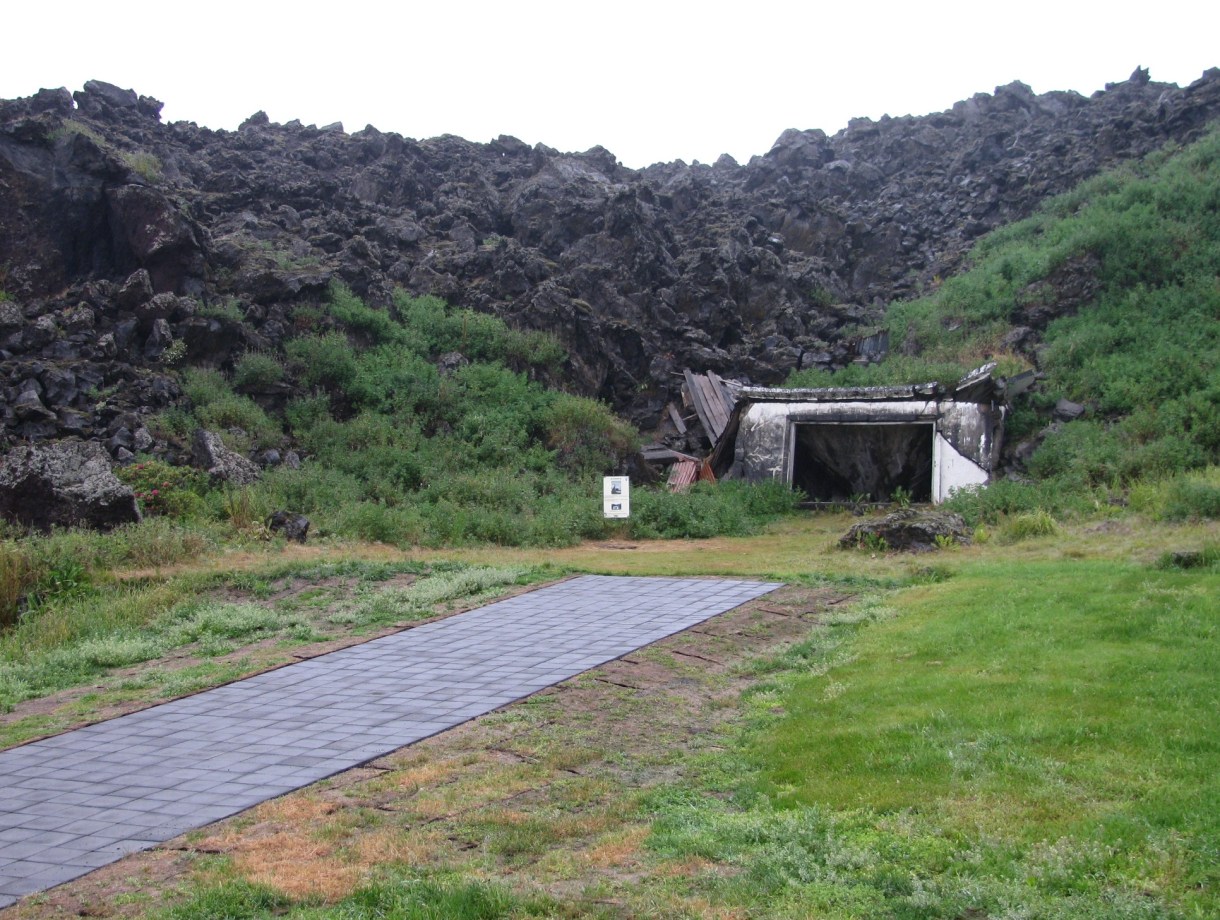 A narrow street that ends in front of a ruined house half buried under jagged boulders.