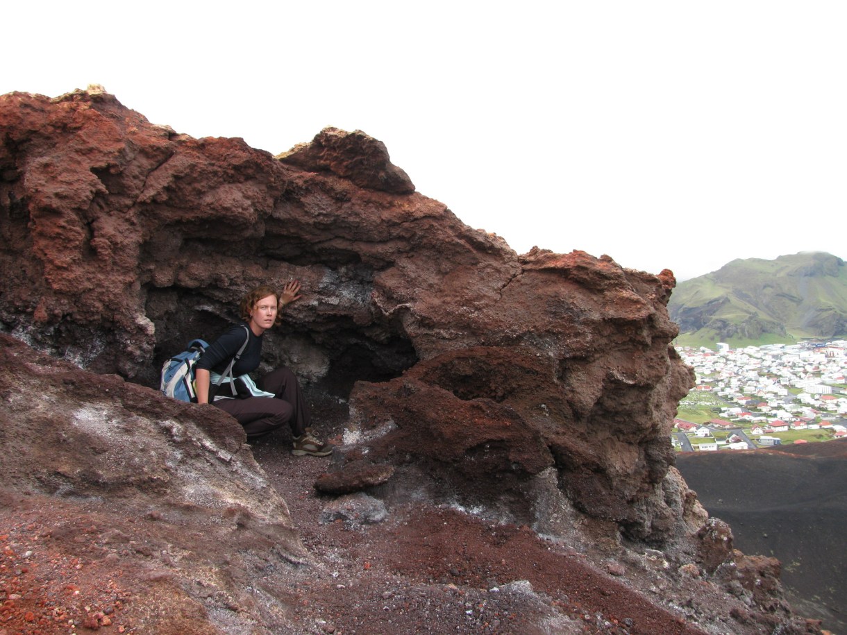 The author is crouching inside a small recess in a large jagged red rock at the top of Eldfell, with her hand extended to touch the side. There is a glimpse of the town and another mountain in the distant background.