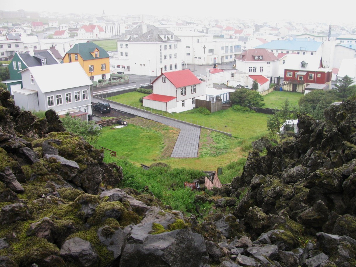 View over Heimaey town showing several white and multicoloured buildings in fog, and a dead-end street ending at a ridge of jagged boulders covered in grass and moss.