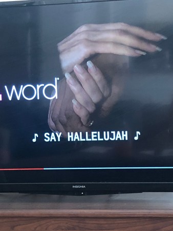 A photo of a freeze frame of The L Work on a flat screen TV sitting on a brown wooden table. It is paused to show a series of hands (with fingernails that are far too long) intertwined. The subtitles show that a song is playing with the lyrics "Say hallelujah."