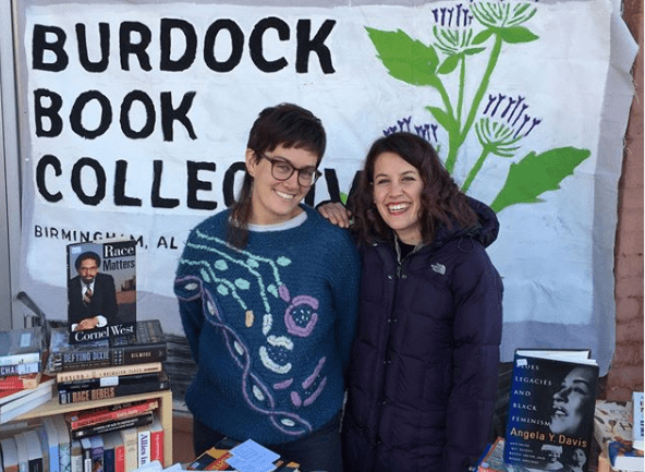 Two people stand in front of a sign reading Burdock Book Collective, smiling at the camera