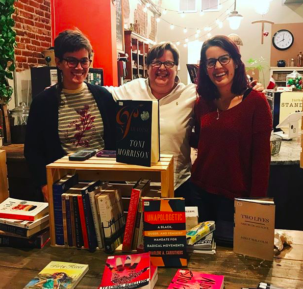 Three people stand behind a display table of books; the person in the center has her arms around the respective shoulders of the two people on either side of her, smiling