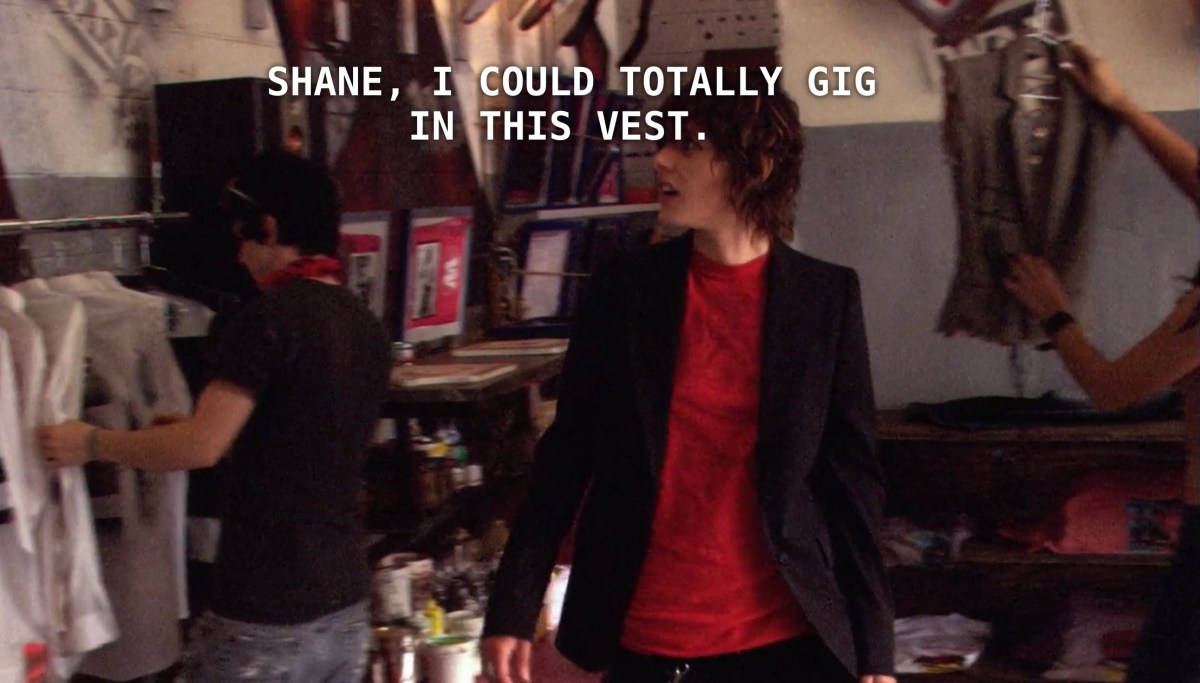 Shane stands in the skate shop, wearing a red t-shirt underneath a black blazer. Carmen is standing behind her. Carmen found a vest and says to Shane, "I could totally gig in this vest."