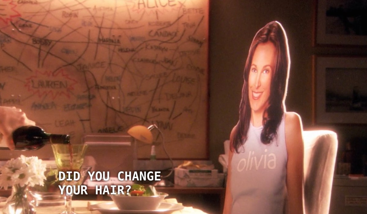 A cardboard cutout of Dana (wearing a purple "Olivia" shirt) sits at a table that is set with wine, flowers, and salad. Alice's massive Our Chart is hanging on the wall in the background. Alice is off screen except for her hand, which is pouring a glass of wine. Alice says, "Did you change your hair?" 