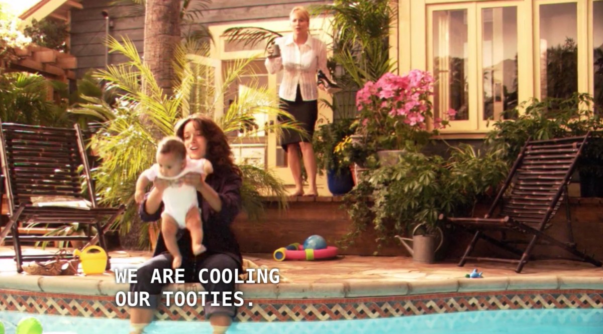 Bette is holding Angie while she sits with her feet dangling over the edge into her backyard pool. Tina stands behind her, wearing a black skirt and white button-up. Bette says to Tina, "we are cooling our tooties."