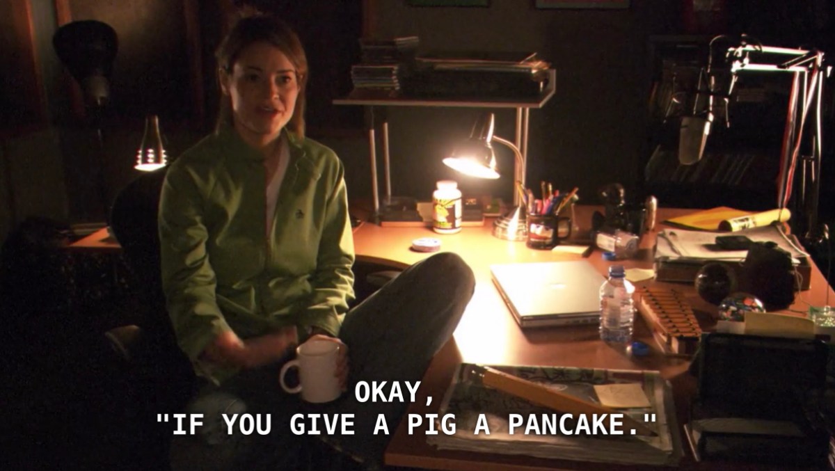 Alice, wearing a green track jacket and holding a white mug, sits at her cluttered desk in the studio. She is pitching ideas to her editor, and just suggested that she read the children's book "If You Give a Pig a Pancake" on air. She goes on to explain that it is her "Earth daughter Angelica's favorite picture book."
