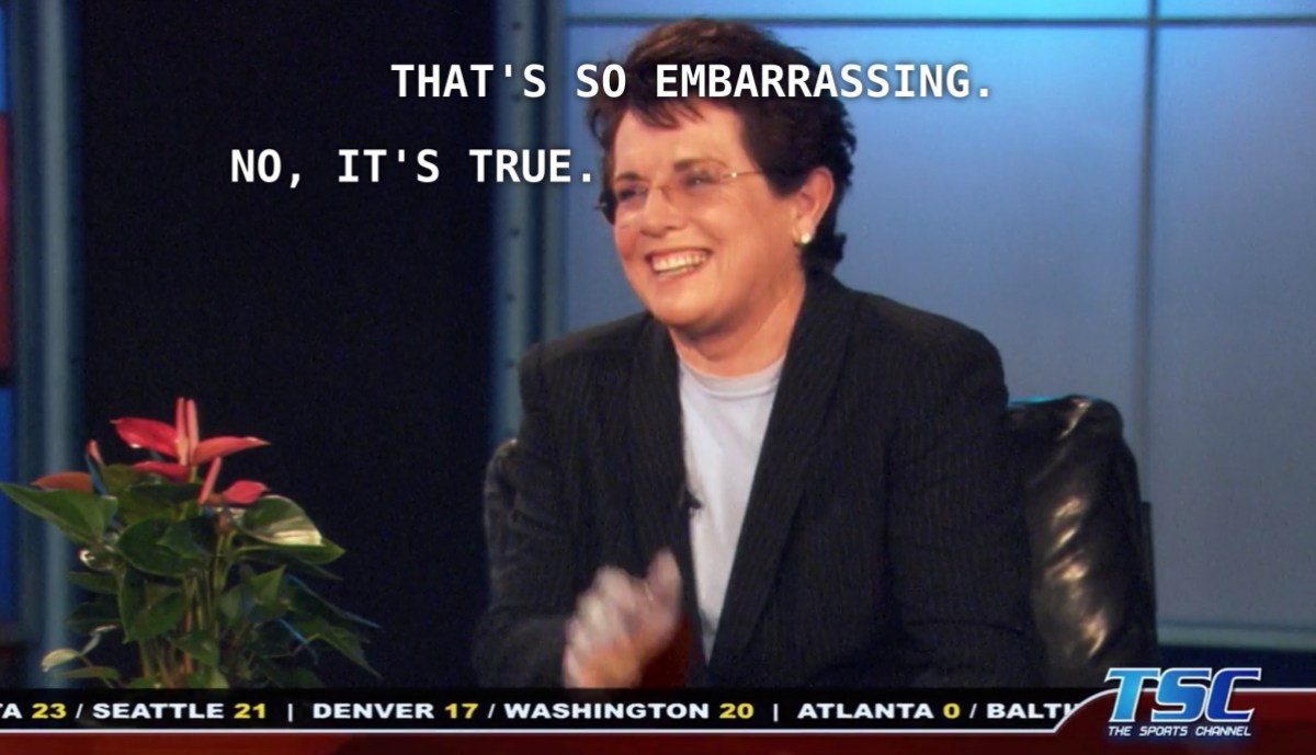 Billie Jean King is wearing a navy blazer and a powder blue shirt and sitting at a desk with a fake-looking plant. She is interviewing Dana for The Sports Channel. Dana has just told her that she is her biggest fan. Billie says, "that's so embarrassing." Dana replies, "no, it's true." 