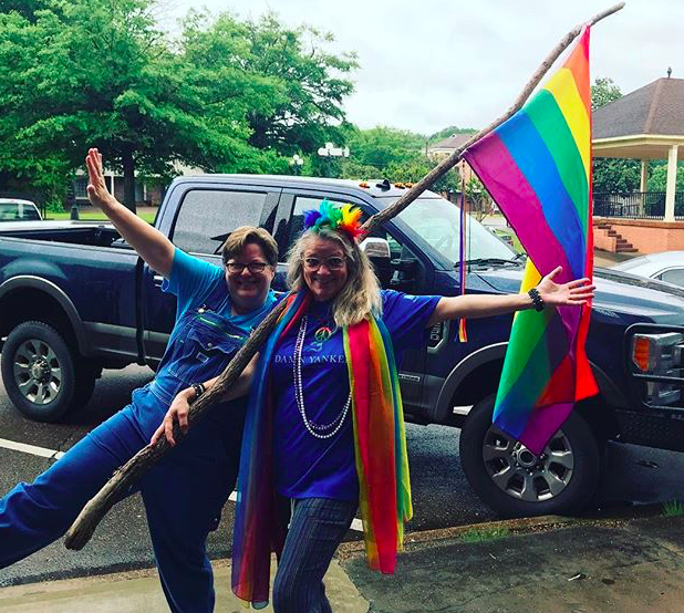 Two people pose with arms spread and wide smiles in a parking lot, holding a rainbow flag and wearing rainbow scarves and feathers