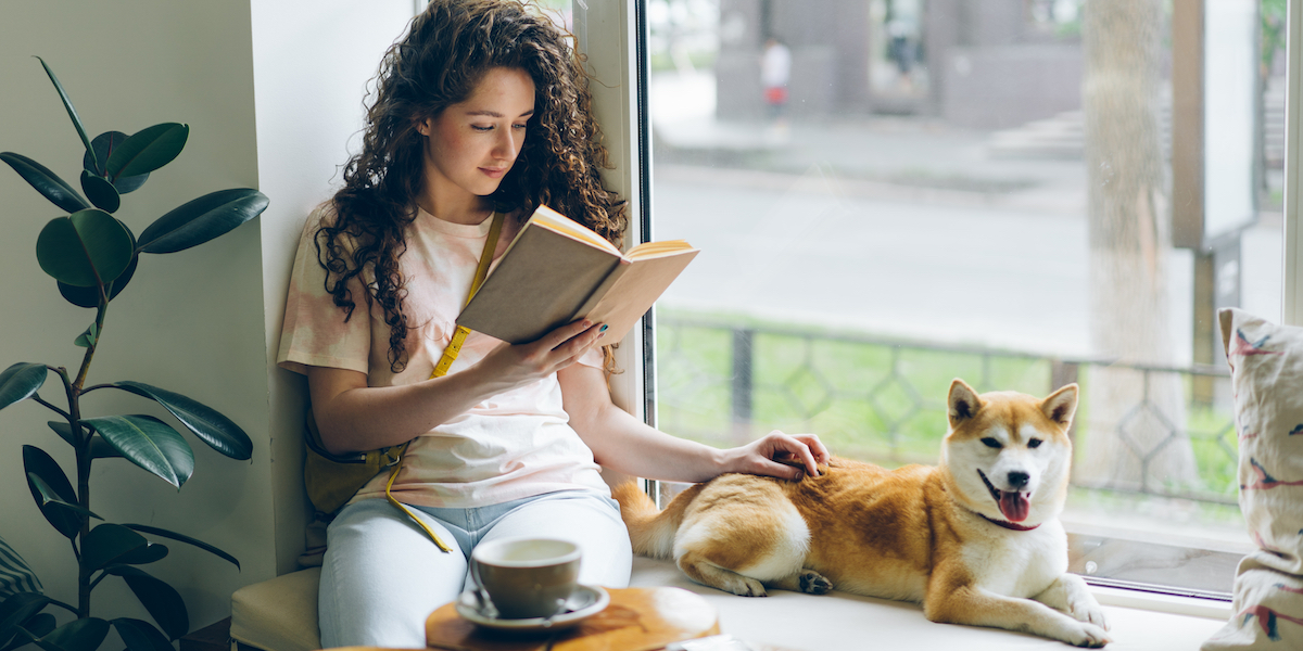 A woman with long curly hair reading in a window, with a tea cup in a saucer and a mid-sized shorthaired dog