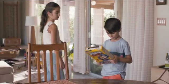 Eli reads the back of a cereal box while his sister Olive walks by.