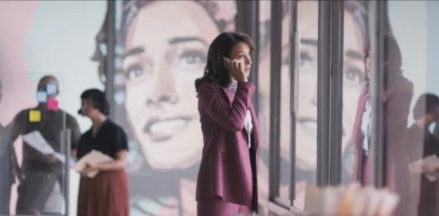 Bette Porter, in a mauve pantsuit is on the phone in her office standing in front of a large painting of Bette