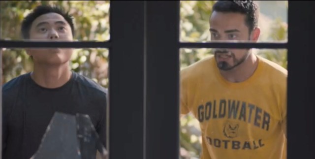 Leo and Jose stand in front of Jose's door. One window pane is broken. Jose is wearing a yellow t-shirt.