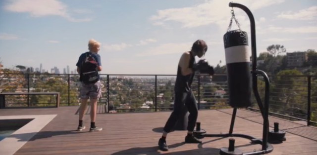 On Shane's deck, Shane punches a punching bag in boxing gloves. Finley, wearing a backpack, looks out at the city.
