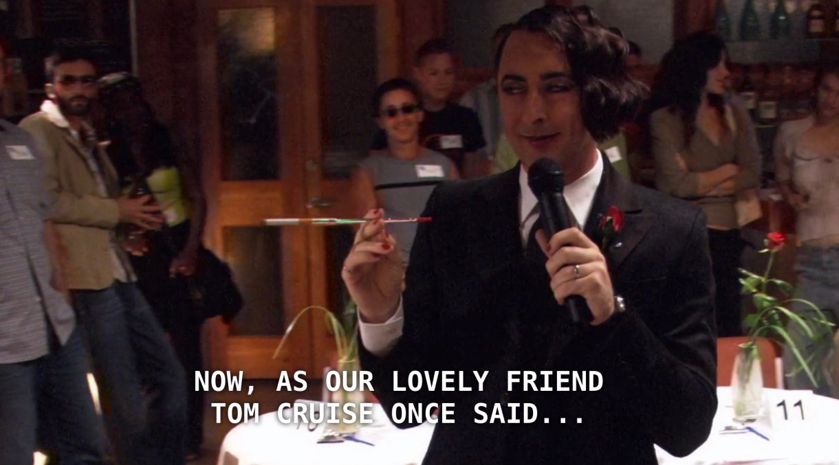 Billie is wearing a black suit, white shirt, black tie, and a monocle, and is holding a microphone in one hand and a long cigarette in the other. He is hosting Bisexual Speed Dating in a crowded room of people. He announces to the crowd, "Now, as our lovely friend Tom Cruise once said, the bar is open!"