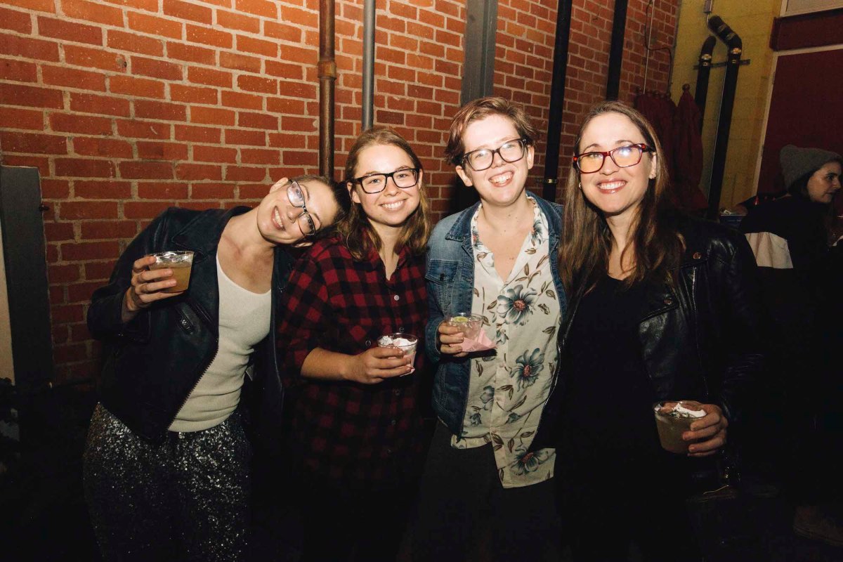 Four people holding drinks pose for a photo against a brick wall inside.
