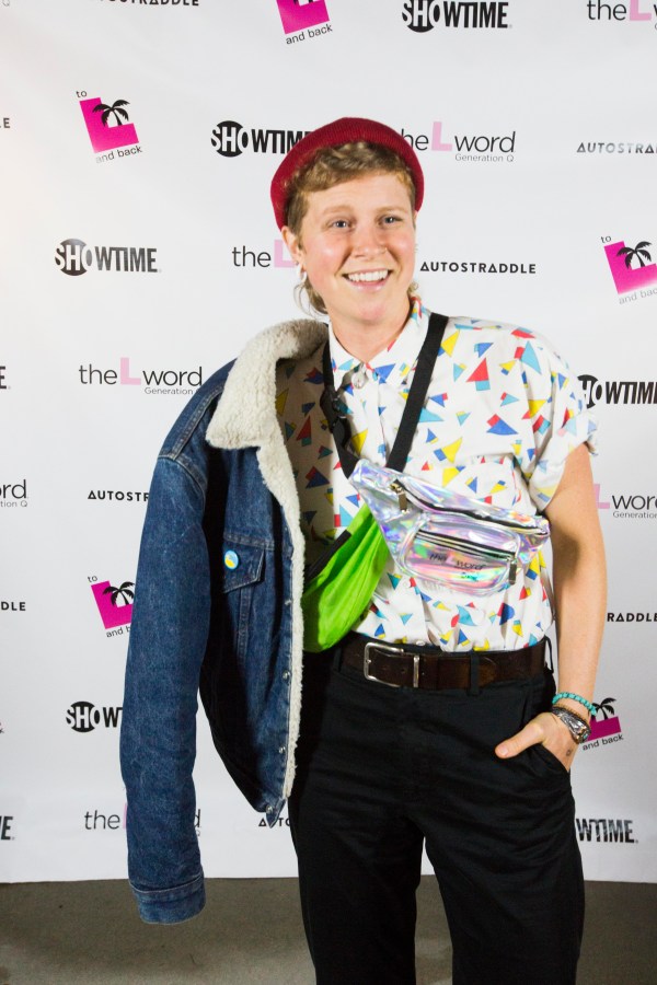 A person posing in front of a promo poster for "The L Word: Generation Q." They are wearing a brightly colored patterned button-up, two fanny packs crossing their chest, and a denim jacket draped over their right shoulder.