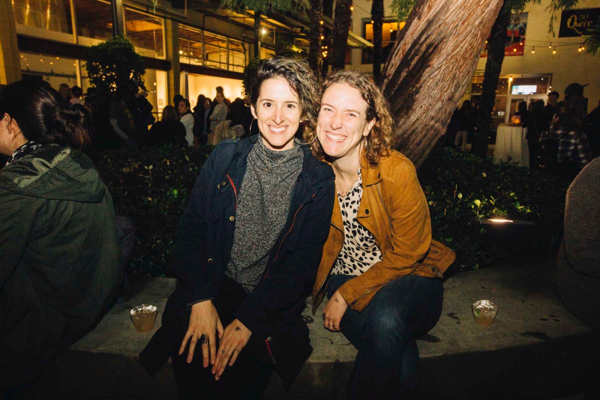 Two people sit outside on a bench and pose for a photo, smiling.