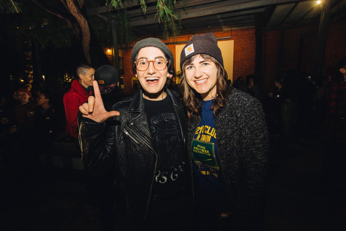 Two people smiling for a photo. The one on the left is wearing a black leather jacket, a grey beanie, and glasses, and is making an "L" with their thumb and pointer finger. The one on the right is wearing a black sweater and a brown beanie with long brown hair.
