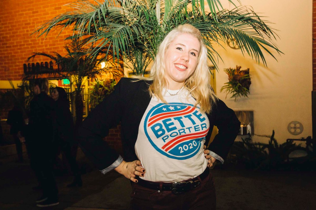 A blonde-haired person wearing a "Bette Porter 2020" t-shirt under a black blazer. They are smiling in front of a palm tree.