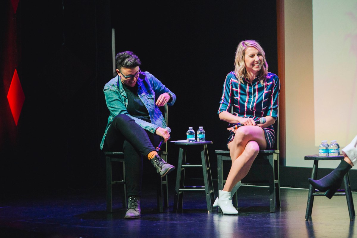 The "To L and Back" hosts Carly Usdin and Riese Bernard sit next to each other on stage. Carly is wearing black jeans and a chambray shirt, and Riese is wearing a blue, red, and white striped dress. 