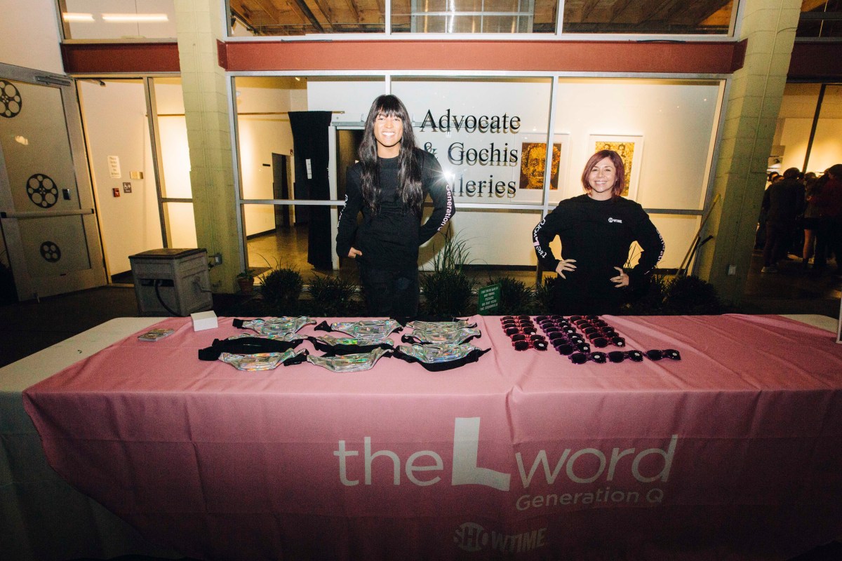 Two people standing behind a merch table with a pink tablecloth with the logo for "The L Word: Generation Q" printed on it. The person on the left is wearing all black, and has long dark hair and bangs. The person on the right is also wearing all black and has short auburn hair. Both are looking at the camera and smiling.