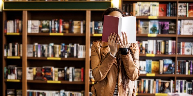 person in a tan jacket in a bookstore holding a book open over their face