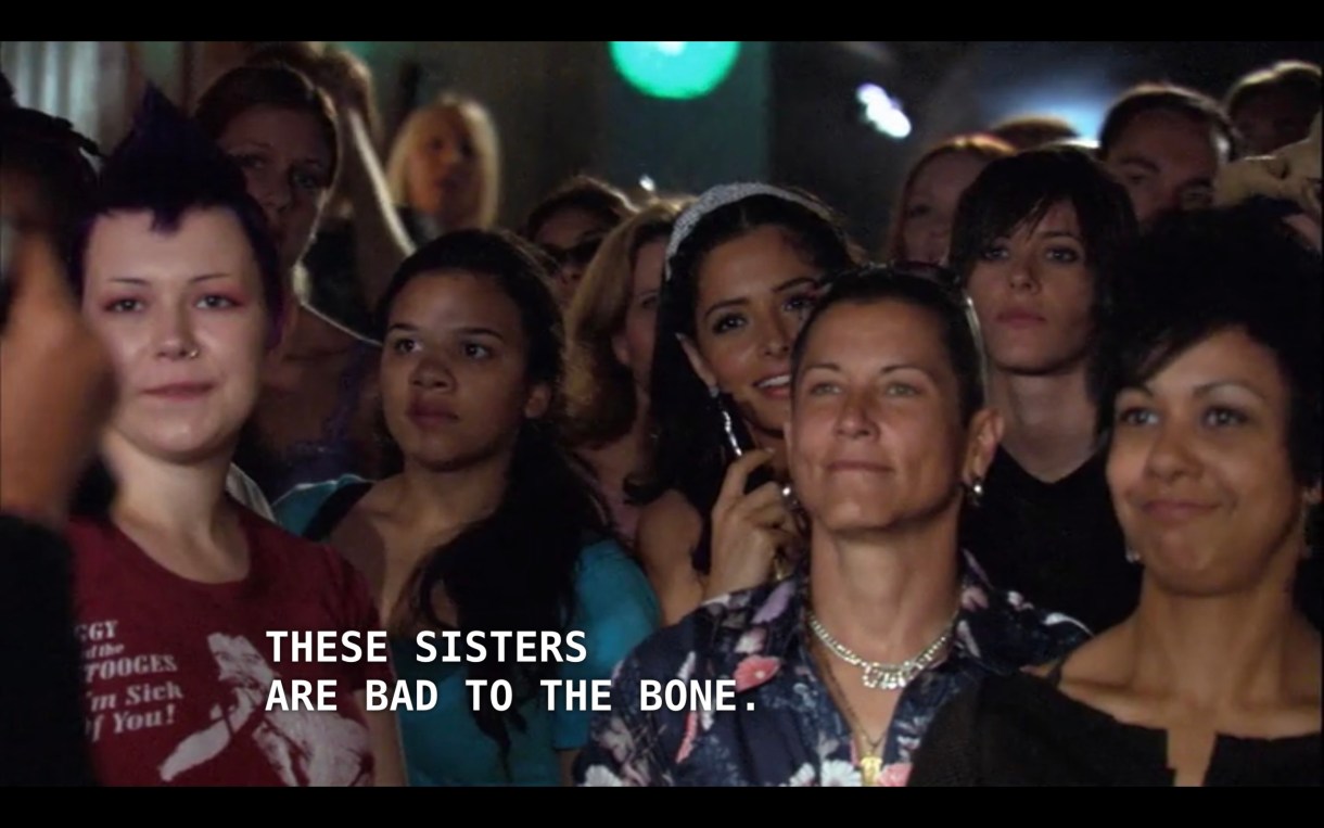 Carmen stands in the crowd, surround by people, at a concert. Off screen, Kit says, "these sisters are bad to the bone."