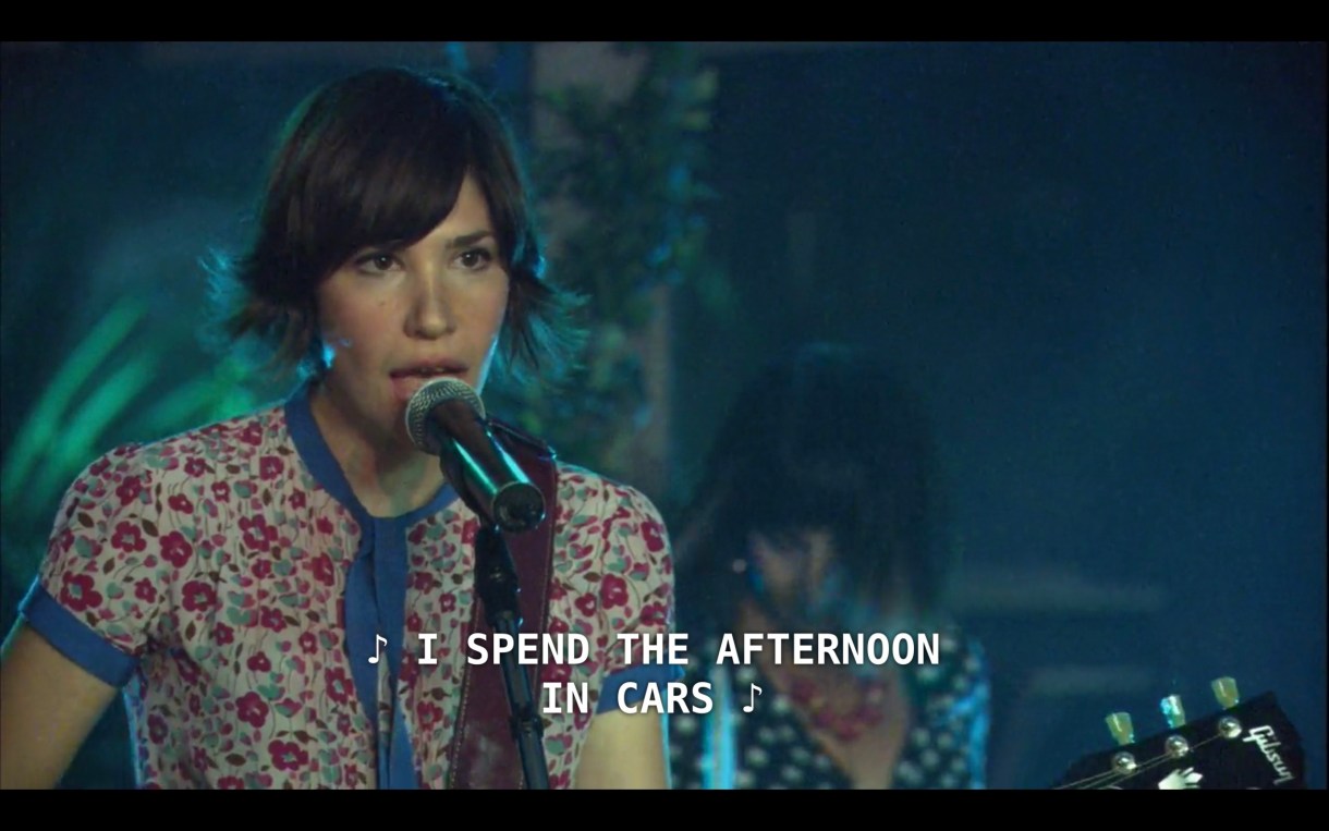 Carrie Brownstein (playing herself in Sleater-Kinney) stands at the microphone on stage performing a concert. She is holding her guitar and has brown short hair that flips out at the ends, and she is wearing a short-sleeve floral shirt. She sings, "I spend the afternoon in cars"