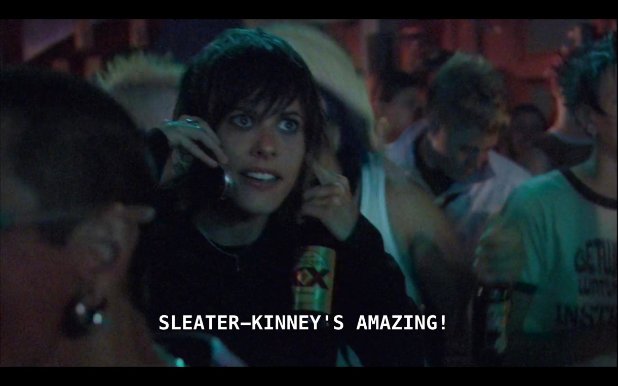 Shane is standing in the crowd at the Sleater-Kinney concert. She is holding a bottle of Dos Equis beer and talking on her cell phone. She says, "Sleater-Kinney's amazing!"