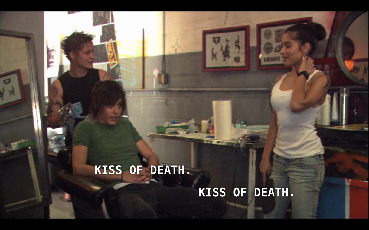 Shane (wearing a green t-shirt) and Carmen (wearing a white tank top and jeans) are in a tattoo shop. Shane is sitting in a chair with a tattoo artist (wearing a black tank top) stands behind her. Shane and Carmen say in unison, "Kiss of death." 