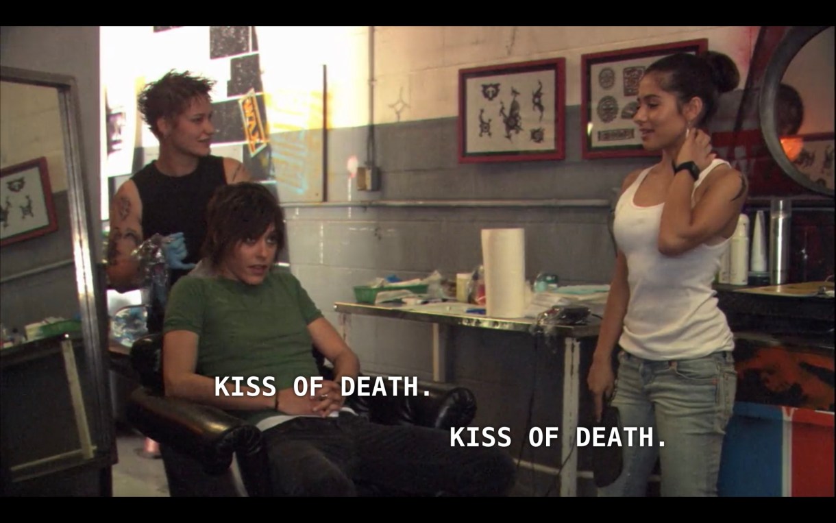 Shane (wearing a green t-shirt) and Carmen (wearing a white tank top and jeans) are in a tattoo shop. Shane is sitting in a chair with a tattoo artist (wearing a black tank top) stands behind her. Shane and Carmen say in unison, "Kiss of death."