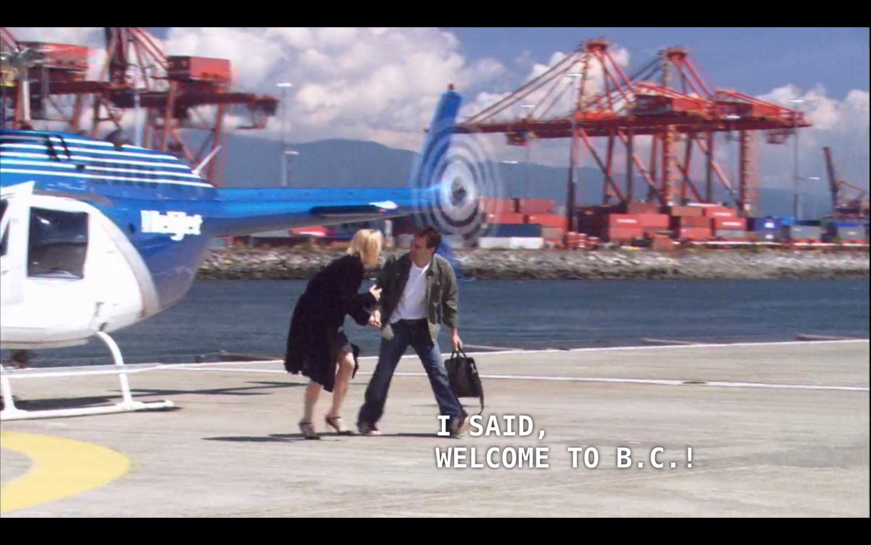 Tina (wearing a black dress and black jacket) has just gotten off a blue and white helicopter. A man (wearing a green jacket, white shirt, and jeans) says to Tina, "I said, welcome to B.C.!"
