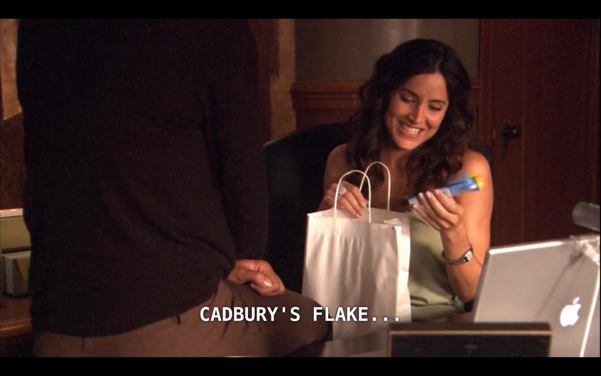 Helena is in her office holding a white paper bag. Dylan is sitting on the desk with her back to the camera, wearing brown pants and a black shirt. Helena has just pulled something out of the bag, smiles, and says, "Cadbury's flake."