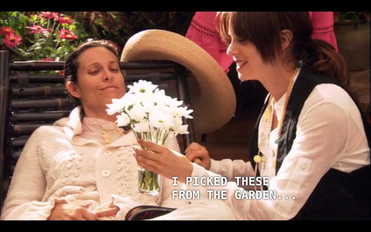 Dana, wearing a white sweater, is laying down on a lawn chair outside. Jenny (wearing a white shirt and black vest) is handing her a bouquet of white flowers. Jenny says, "I picked these from the garden"