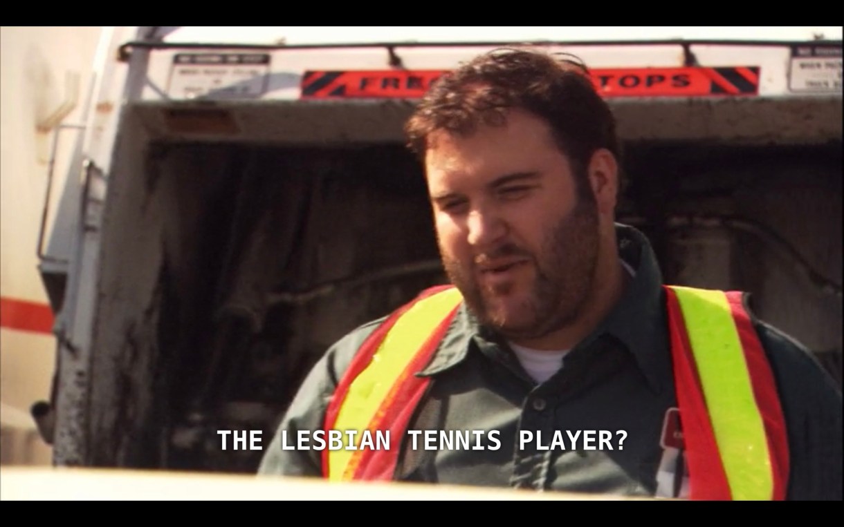 A sanitation worker (wearing a dark green shirt and neon safety vest) stands in front of a garbage truck. He says to Alice (who is off screen), "The lesbian tennis player?" when Alice inquires about the life-size Dana that she threw away.