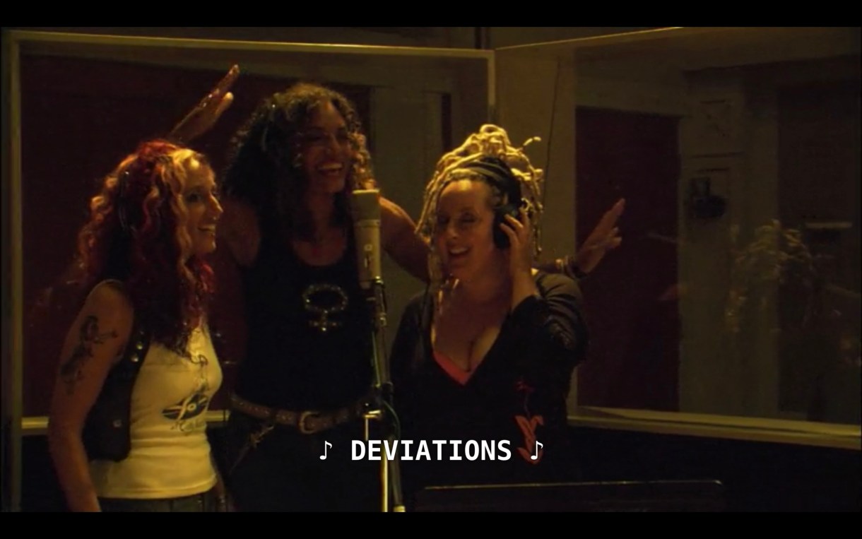 The 3 band members of Betty are standing around a microphone in a recording studio. The subtitles show that they are singing the lyric "Deviations."