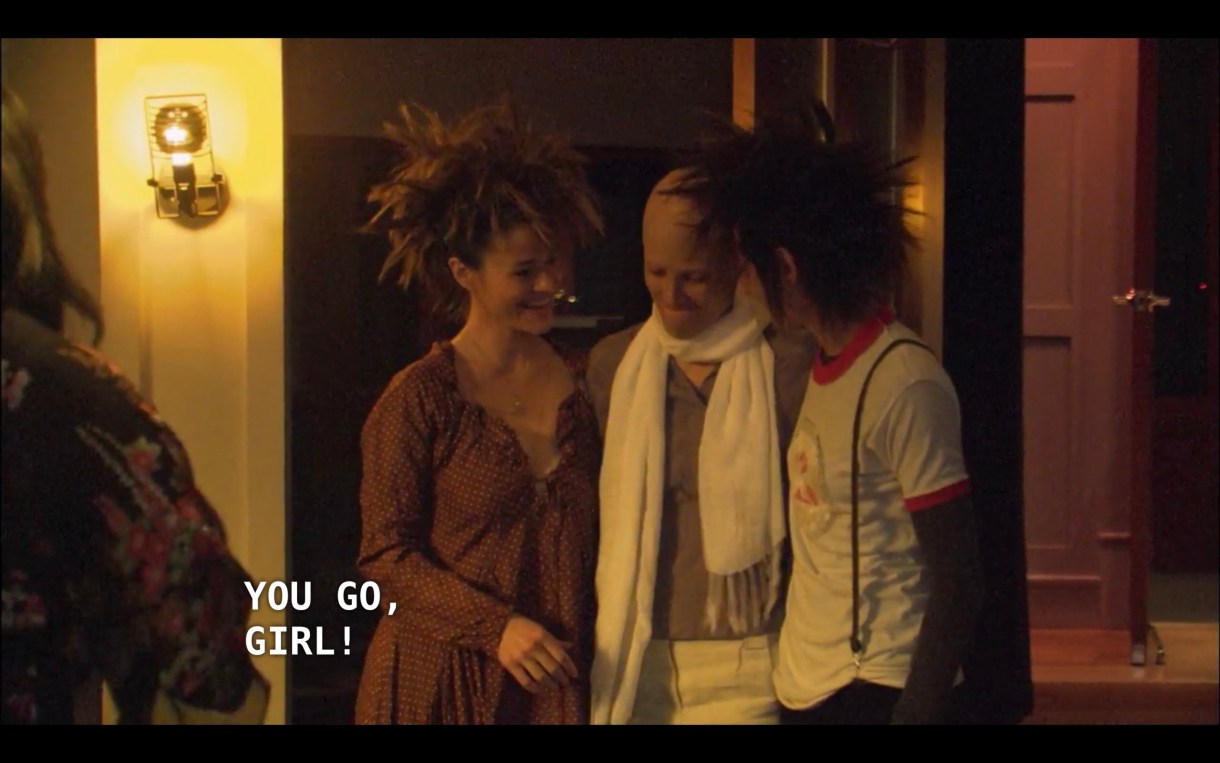 Alice and Shane (both wearing short spiky wigs) stand on either side of Dana, who is now bald and wearing a chunky white scarf around her neck. They scream, "You go, girl!"