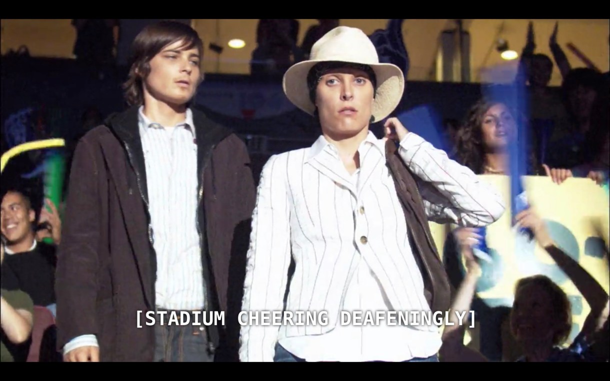 Max and Dana (who is wearing a large white fedora, mind you) stand in a crowded stadium bleachers. The subtitles read "[stadium cheering deafeningly]"