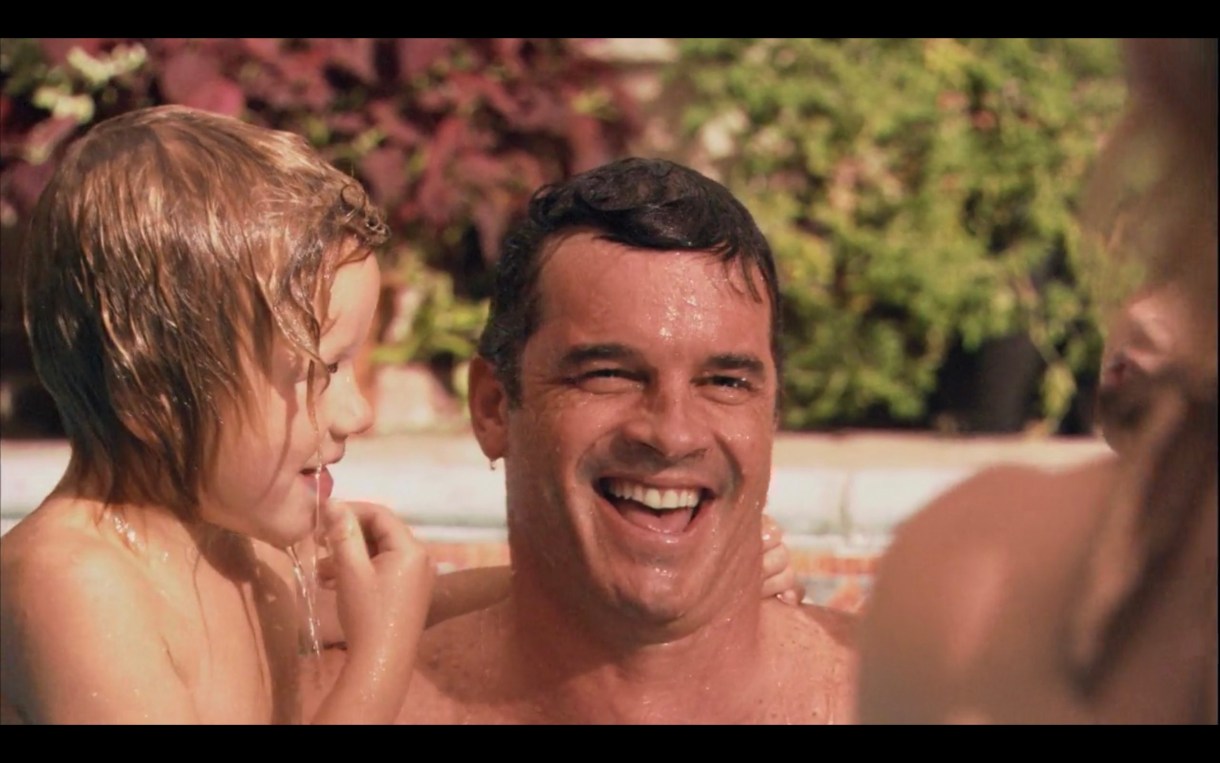 Henry is holding one of his blonde-haired sons in the pool during the kids' swim lesson. They are both wet and Henry has a goofy smile on his face.