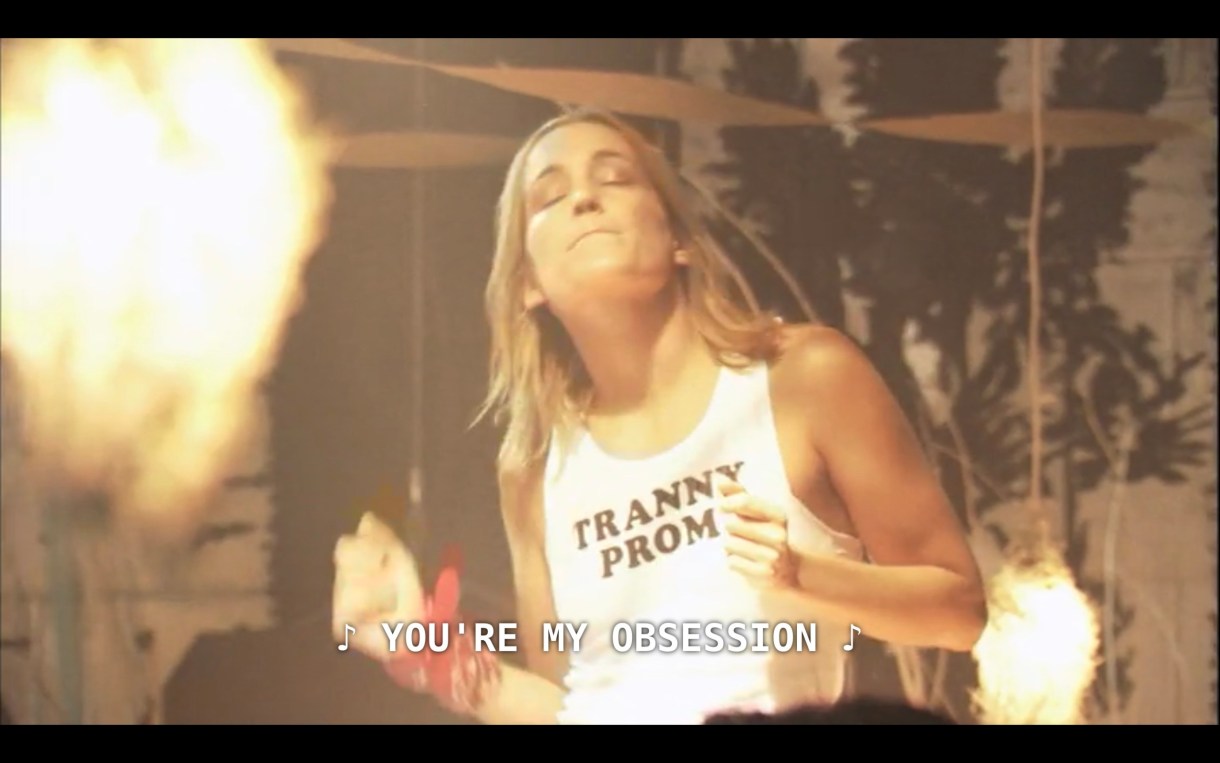 A person with long blonde hair is dancing at the queer prom event. They are wearing a white tank top that says "Tranny Prom" in black letters.