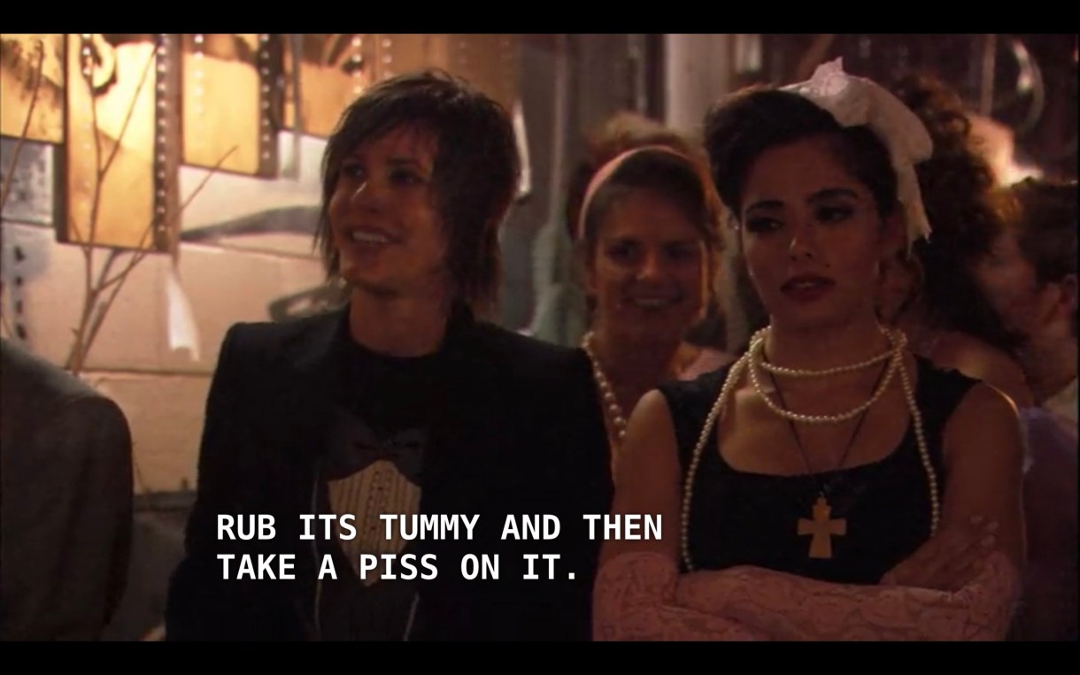 Shane (wearing a classic black tee under a black blazer) is standing next to Carmen (wearing a black dress, pink lace gloves, pearls, and a white bow in her hair) at the Queer Prom. Shane tells Carmen that Carmen's mom told her about how when Carmen was little and has a teddy bear that she would "rub its tummy and then take a piss on it."