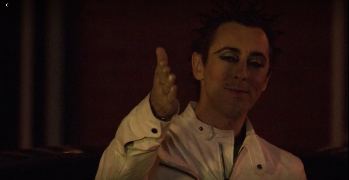 Billie is wearing eyeliner and a white leather motorcycle jacket. He has one hand outstretched in a "tada!" gesture.