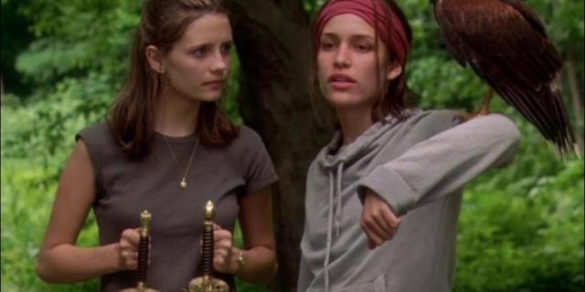 A film still from Lost & Delirious where one girl stands on the left holding two swords by the hilt and looks quizzically at the girl on her right, who's wearing a red sweatband over her forehead and hoisting a falcon on her arm