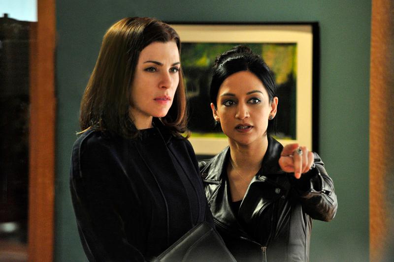 Image: Alicia Florrick, played by Juliana Marguiles, is wearing a black shirt and cardigan and holding a legal brief. She is standing next to Kalinda Sharma, played by Archie Panjabi, who is wearing a black leather jacket and pointing towards the camera. Alicia is looking in the direction that Kalinda is pointing. Behind them is a green wall with a painting hanging on it.