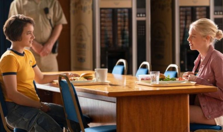Image: Jules, in a yellow t-shirt, sits across a table from Hanna. Both have food trays in front of them. There is a guard standing behind the table, just his torso is visible.