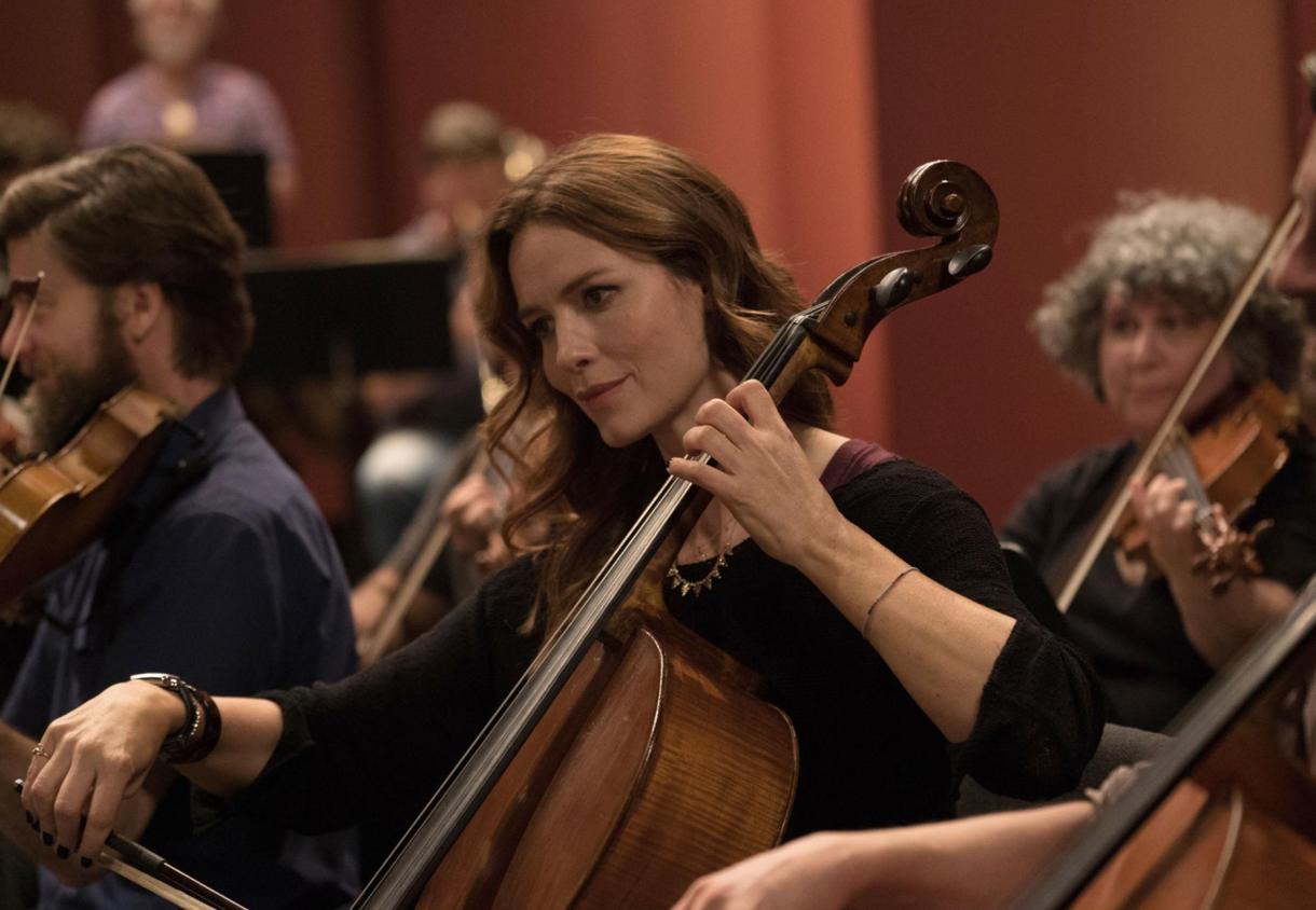 Image: a white woman with long flowy hair in a symphony playing the cello