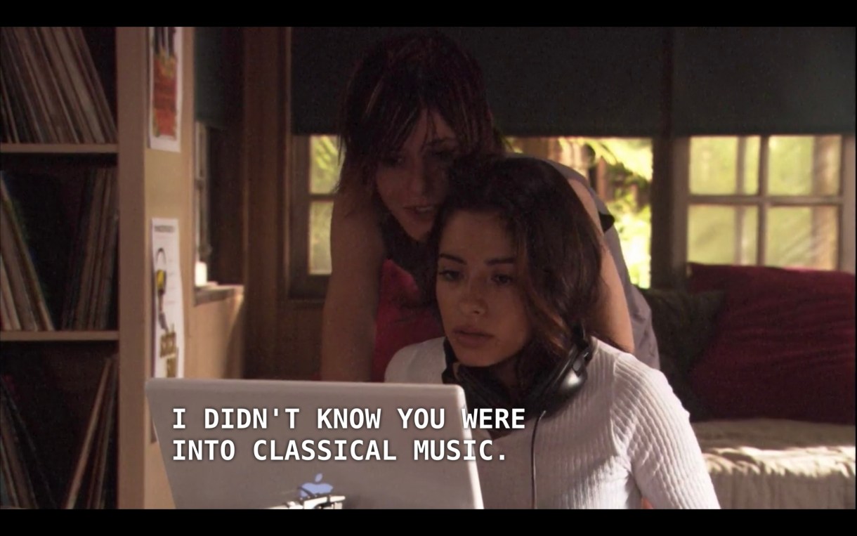 Carmen is sitting with her computer at a table in Shane and Jenny's house. She is wearing a white sweater and her headphones are hanging around her neck. Shane is looking over her shoulder and says, "I didn't know you were into classical music."