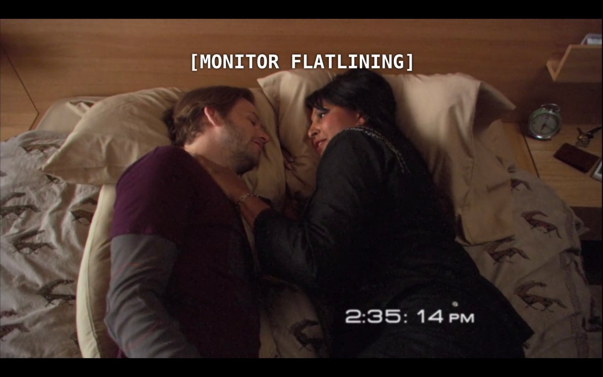 Angus (wearing a maroon t-shirt over a grey long sleeve shirt) and Kit (wearing all black) lay in bed facing each other. The subtitles read, "[Monitor flatlining]" referring to what is happening in Dana's hospital room at the same moment.